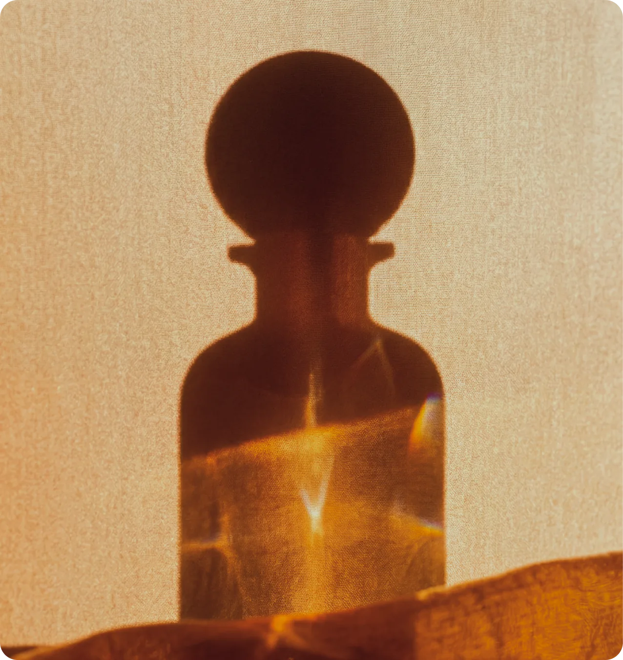 Alma di Alba — a bottle silhouette diffused through warm amber fabric, sunlight catching the spherical cap in refracted color