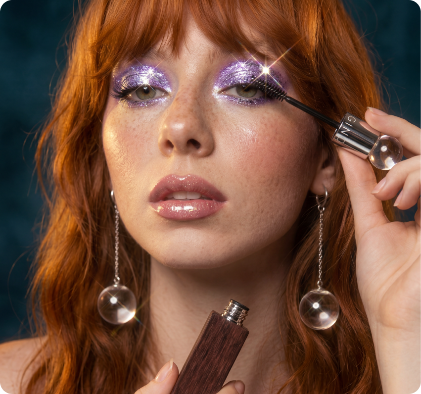 Ginga — a red-haired model with lavender glitter eyeshadow applying Ginga mascara, wearing chrome-orb earrings against a teal backdrop