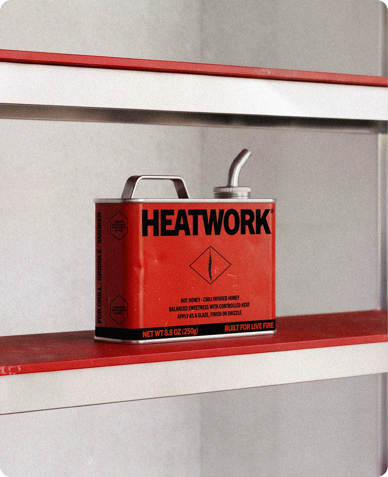 Heatwork: the red tin standing on a red shelf rail against a pale wall, its spout angled out, the HEATWORK wordmark and the diamond mark facing forward