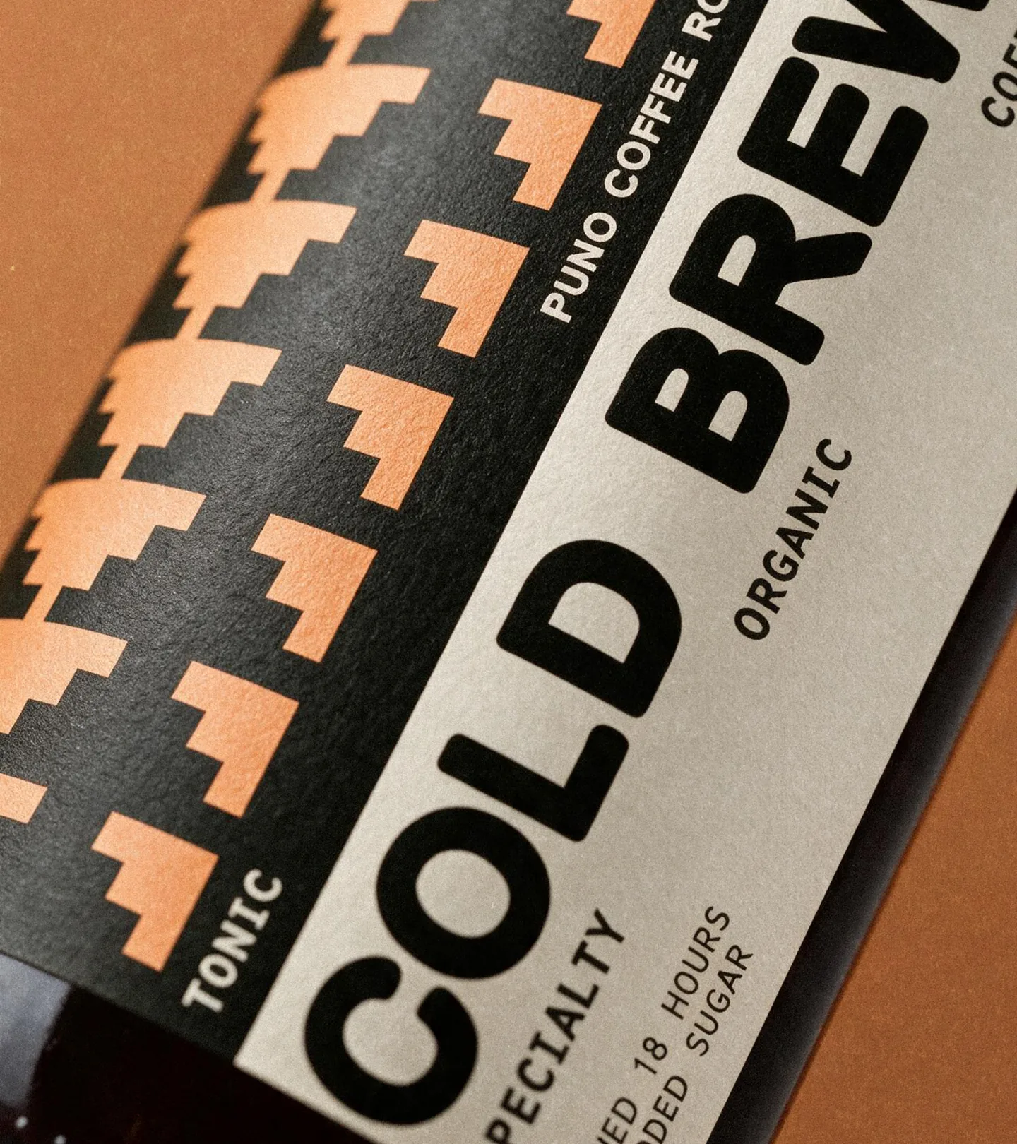 Puno — extreme close-up of a cold brew bottle label on a terracotta backdrop, cream and black with the peach-and-black pyramid pattern down the side and 'PUNO COFFEE ROASTERS · SPECIALTY · ORGANIC · AGED 18 HOURS · NO ADDED SUGAR' running across the label