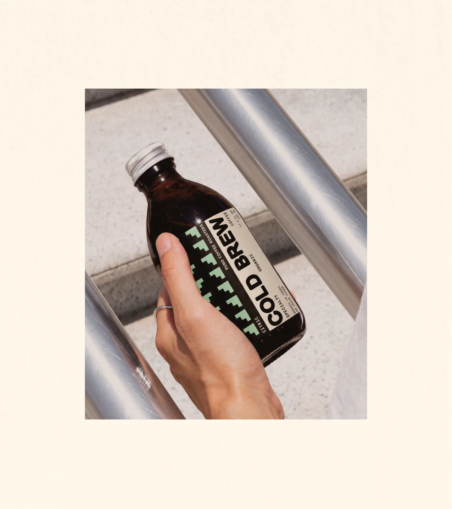 Puno — a ringed hand holding an amber 'CITRIC / COLD BREW' Puno bottle labeled 'PUNO COFFEE ROASTERS · SPECIALTY · ORGANIC · BREWED 18 HOURS · NO ADDED SUGAR' against a metallic handrail and concrete step