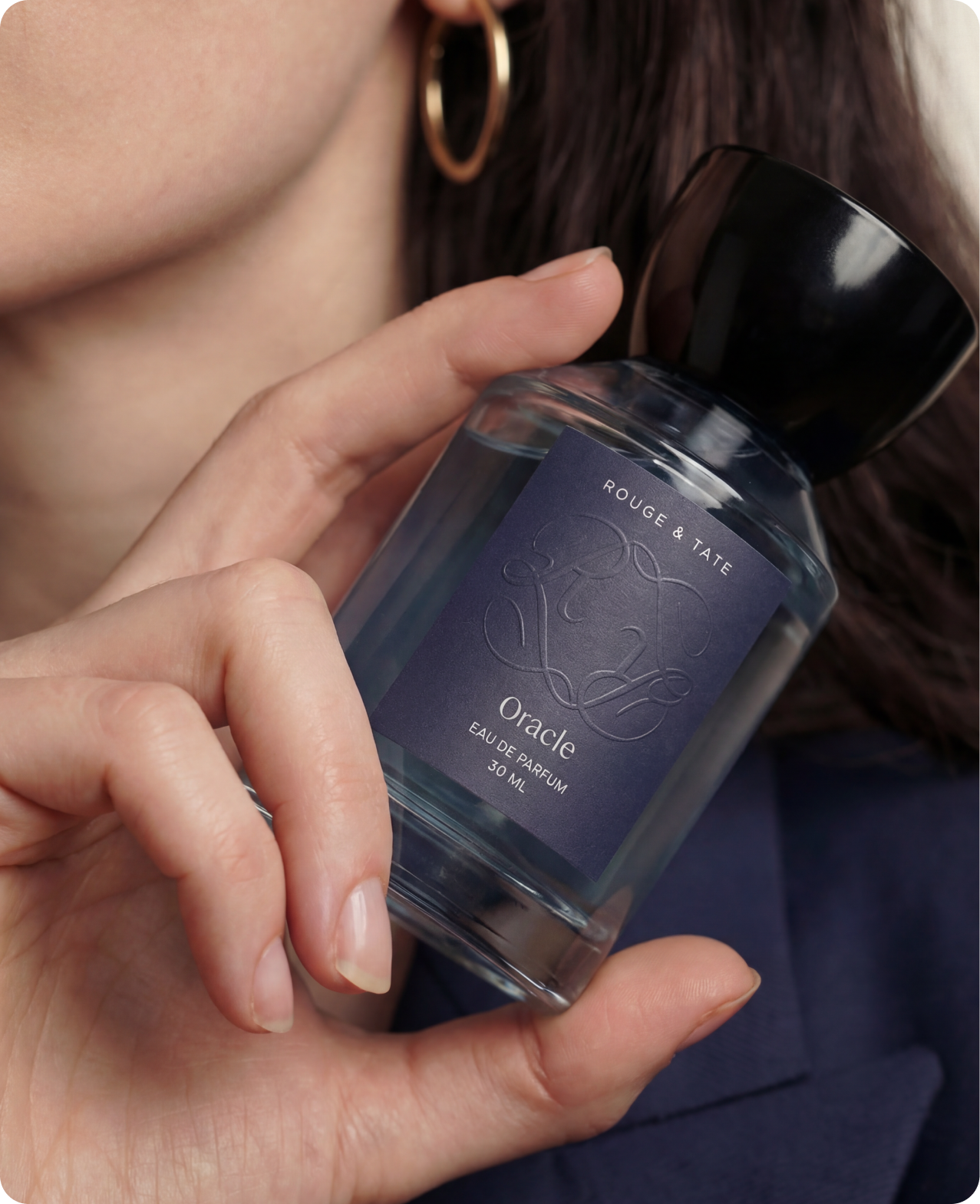 Rouge & Tate — closeup on the Oracle 30 ml bottle held near the model's hoop earring, cool blue label in soft focus