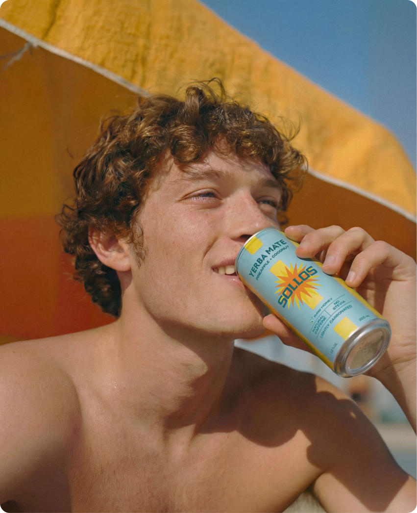 Sollos — a young man drinking a Sollos yerba mate can under an orange beach umbrella in bright sun