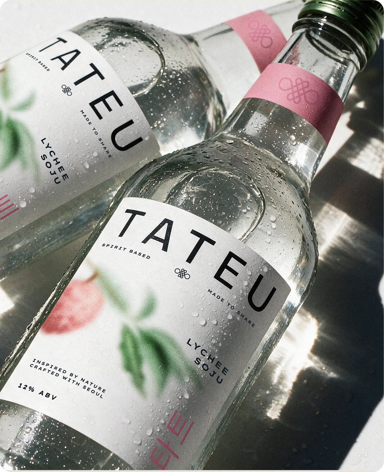 Tateu — close-up of two chilled bottles beaded with condensation, the front label reading TATEU above 'SPIRIT BASED' and 'MADE TO SHARE' with LYCHEE SOJU set vertically, a soft-focus lychee branch illustration, Hangul characters and 12% ABV