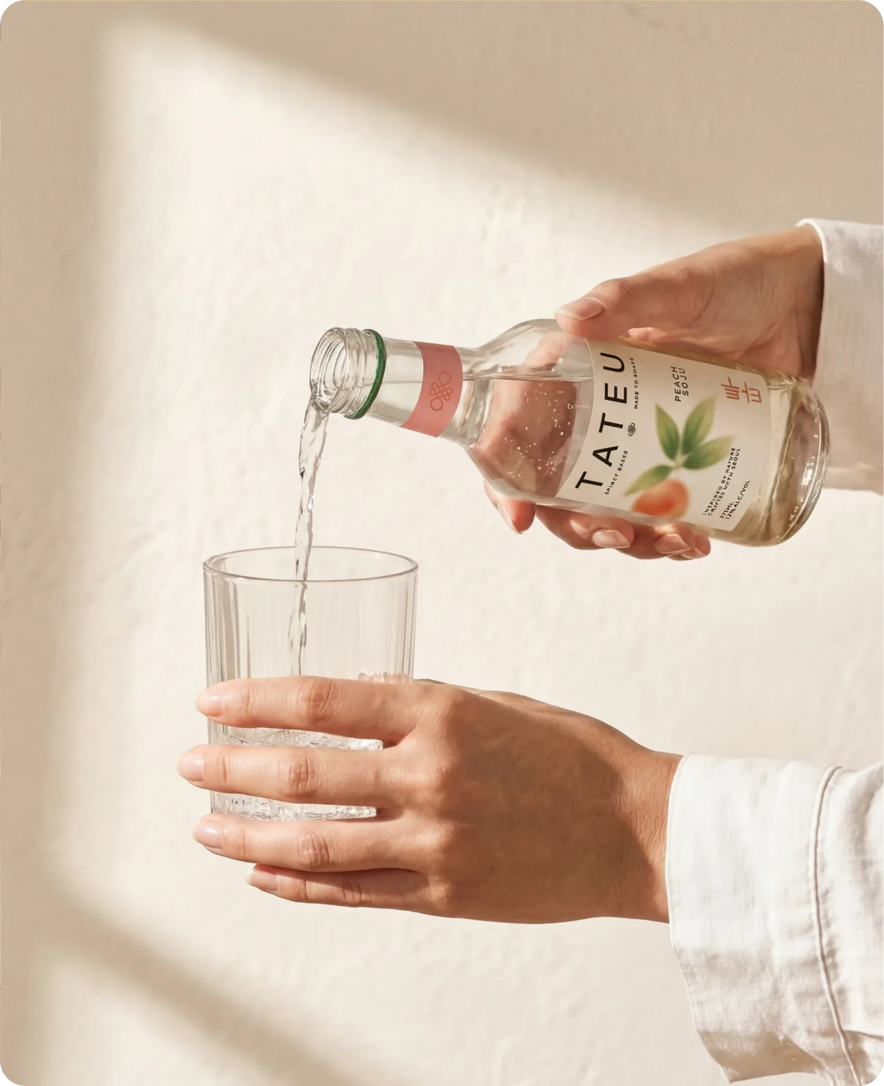 Tateu — a hand pouring Peach Soju from a Tateu bottle into a fluted glass held in the other hand, shot against a warm cream wall with a soft diagonal shadow