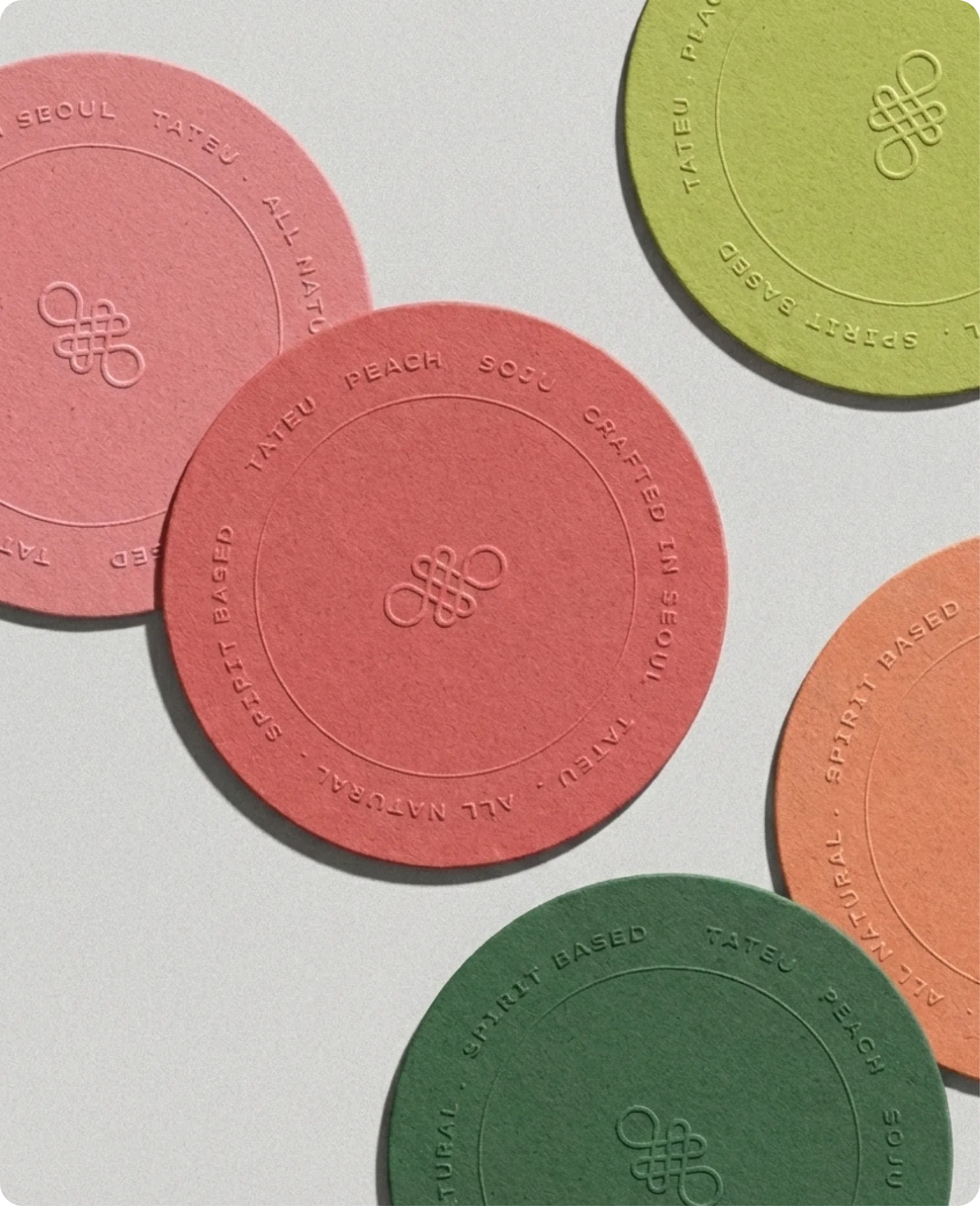 Tateu — embossed pulp-board coasters scattered on a pale surface in the flavour palette of dusty pink, olive, forest green and terracotta, each ringed with 'SEOUL · TATEU · ALL NIGHT · PEACH SOJU · CRAFTED IN SEOUL · SPIRIT BASED' around the central monogram