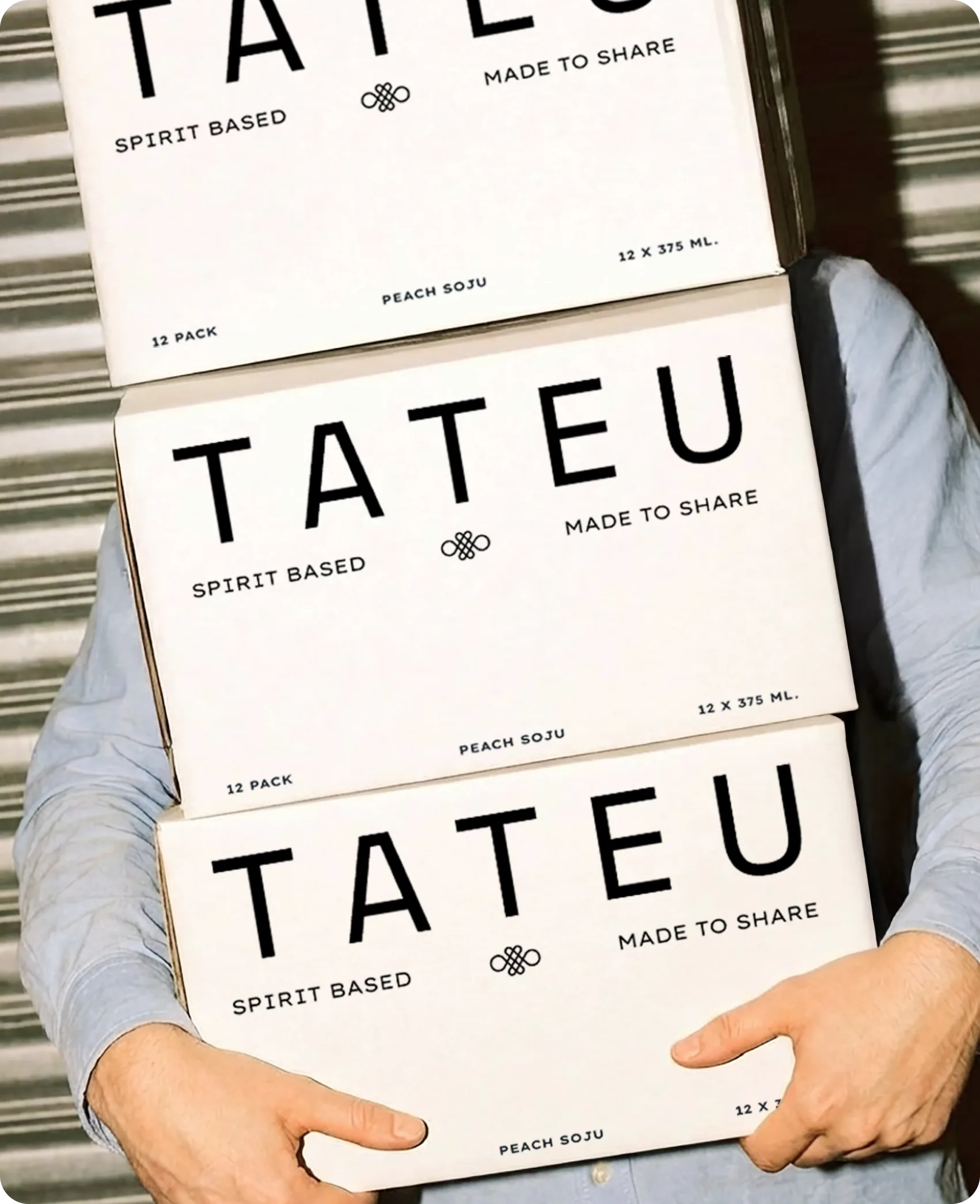 Tateu — a person carrying a stack of three cream shipping cases printed with the TATEU wordmark, 'SPIRIT BASED', 'MADE TO SHARE' and '12 PACK · PEACH SOJU · 12 X 375 ML'