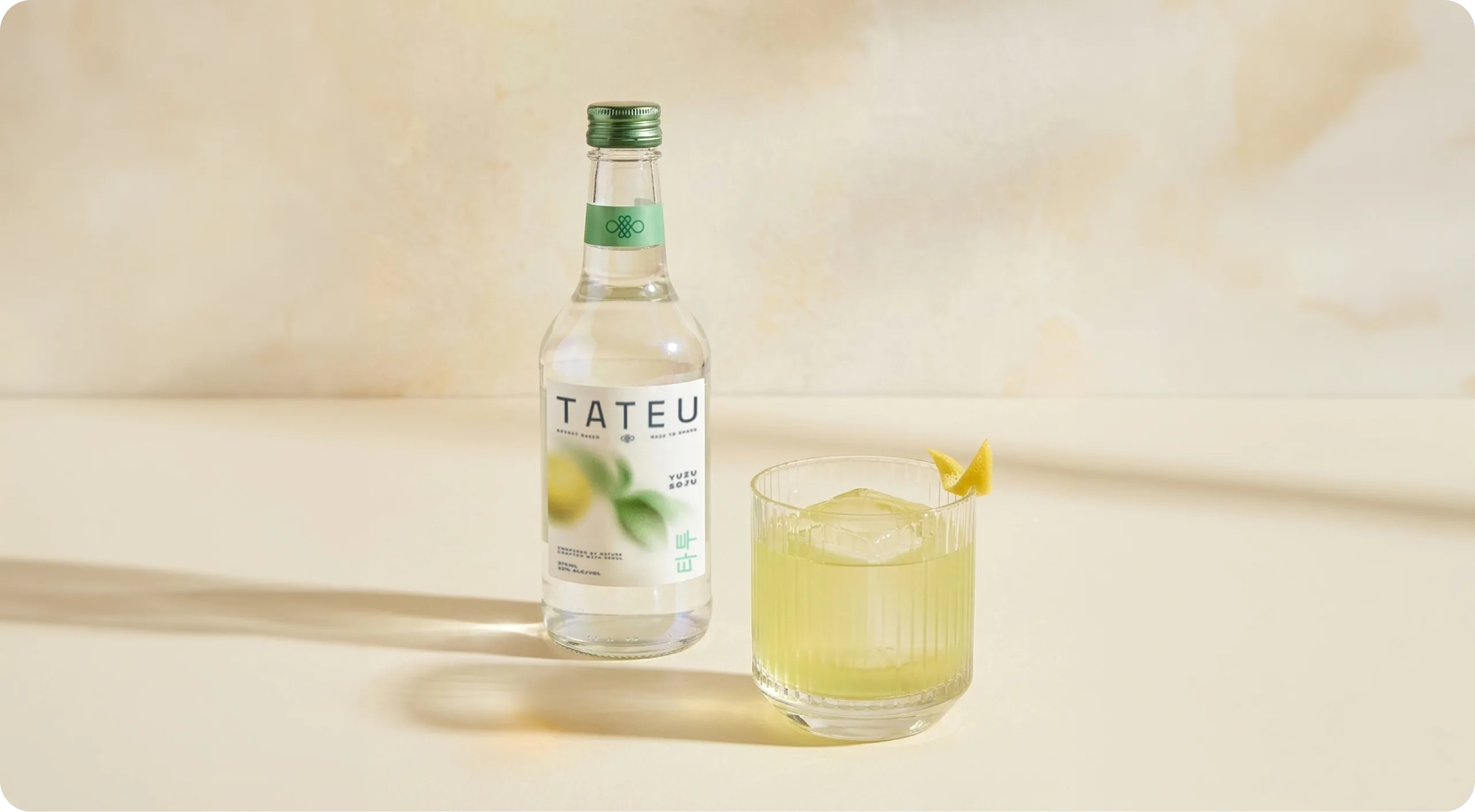 Tateu — a bottle of Yuzu Soju beside a fluted rocks glass of the served cocktail over ice with a citrus twist, both casting long shadows across a warm cream set
