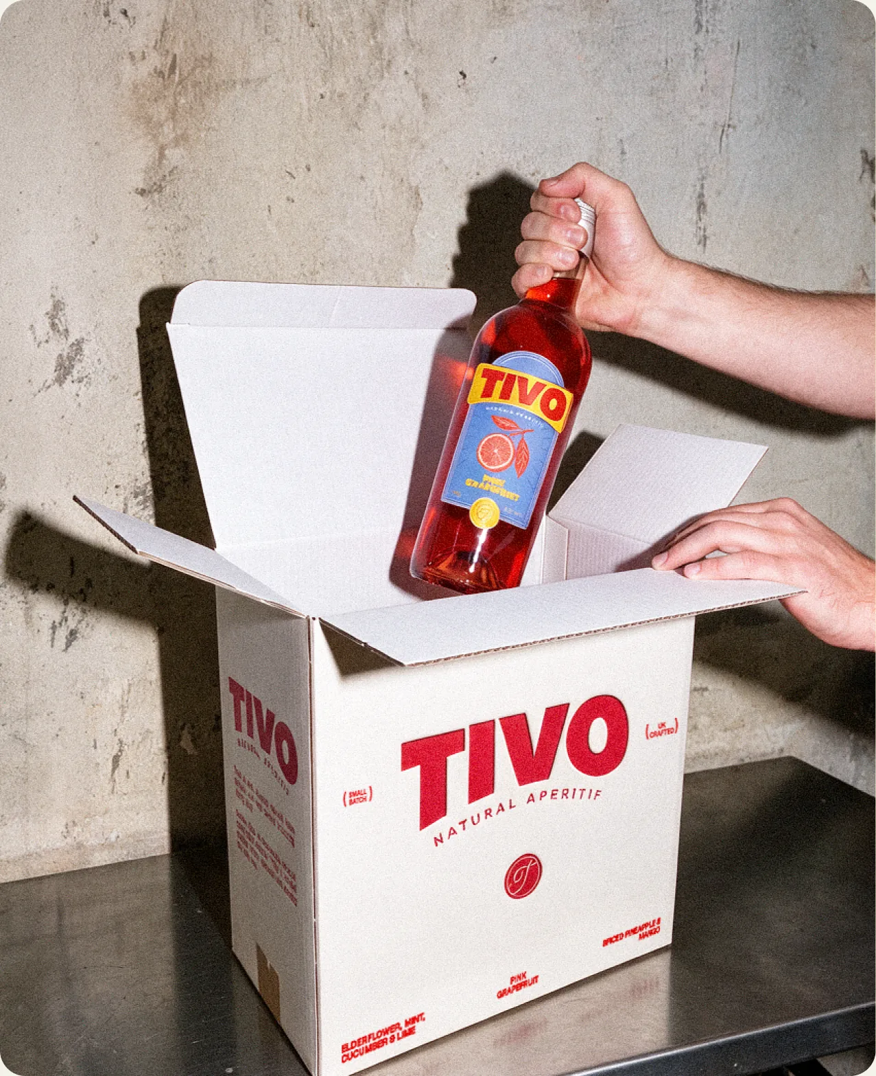 Tivo: two hands lifting a Pink Grapefruit bottle out of an open white shipping box printed with the Tivo wordmark, the words natural aperitif and the cursive T seal, on a concrete floor