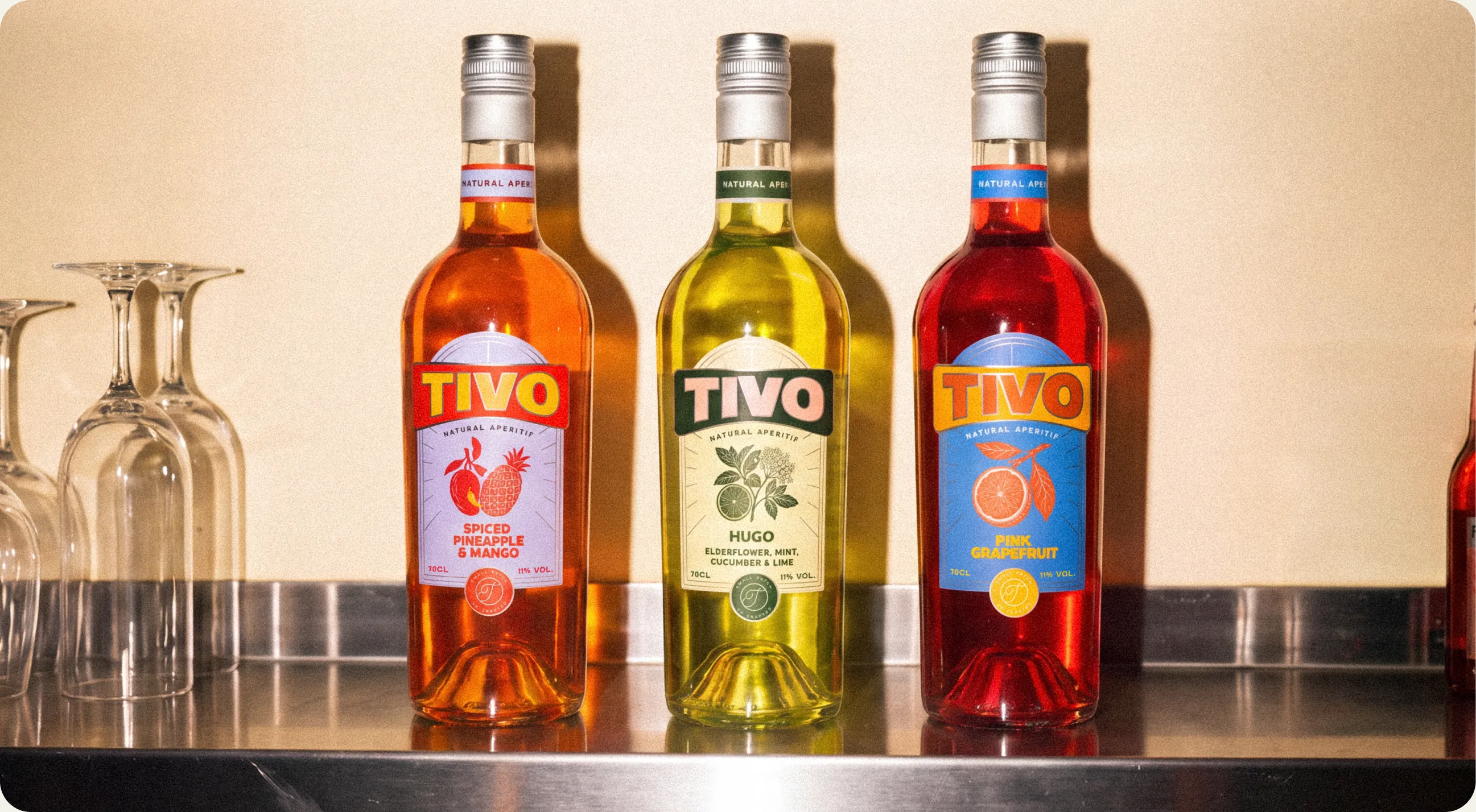 Tivo: the three bottles standing in a row on a stainless bar shelf beside upturned wine glasses, Spiced Pineapple and Mango, Hugo, and Pink Grapefruit